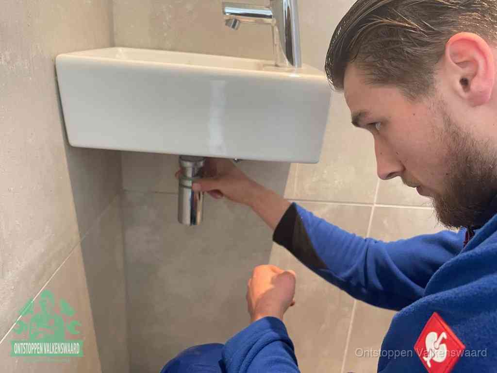 Een monteur draait met de hand de zwanenhalssifon onder een fonteinwasbak in de toiletruimte los om de bron van rioollucht in Valkenswaard te vinden