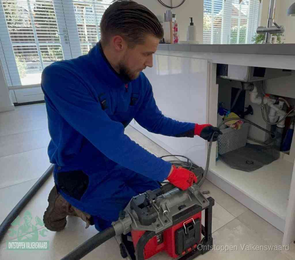 Een monteur met rode handschoenen knielt in een keuken en voert een elektrische ontstoppingsveer in tijdens het professioneel afvoer ontstoppen in Valkenswaard