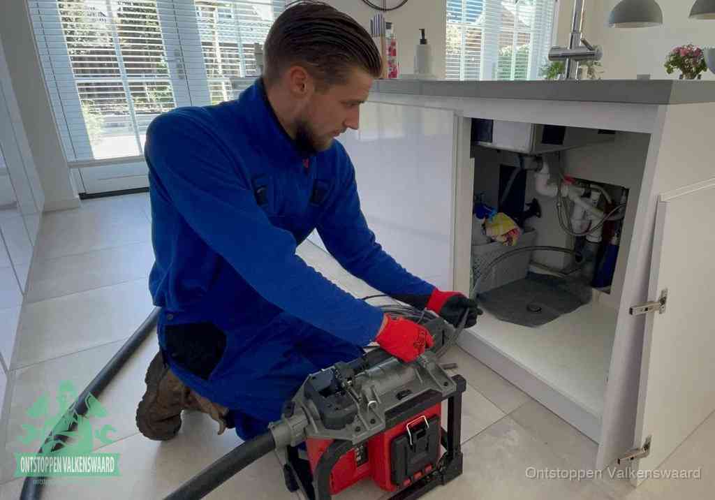 Een monteur voert een professionele elektrische ontstoppingsmachine in de afvoer onder de spoelbak tijdens het gootsteen ontstoppen in Valkenswaard